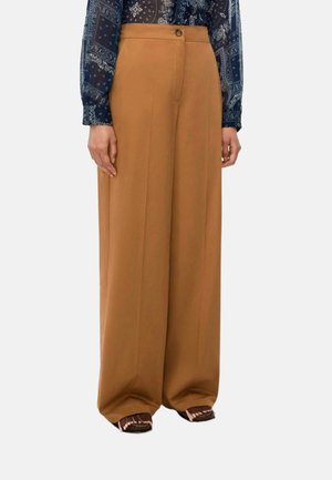 Donna che indossa pantaloni color cammello a vita alta e gamba larga abbinati a una camicetta blu scuro con motivo e sandali marroni.