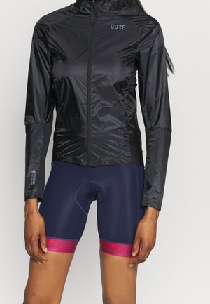 Close-up van een persoon die een zwart Gore hardloopjack met rits en marineblauwe fietsshorts met roze banden draagt.