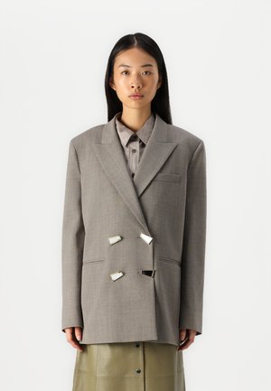Blazer grigio doppiopetto con ampi rever, una tasca sul petto e dettagli triangolari metallici, abbinato a una camicia a righe sotto.