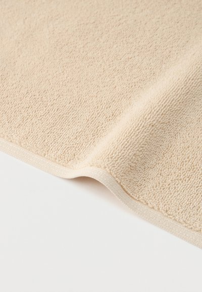 Κοντινό πλάνο πετσέτας από μπεζ ύφασμα τέρρυ που δείχνει την υφή με θηλιές και την διπλωμένη άκρη της σε λευκή επιφάνεια.