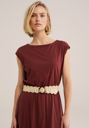 Žena sa kratkom plavom kosom u bezrukoj bordo haljini i bež tkanom remenu s okruglom kopčom, stoji ispred neutralne pozadine.