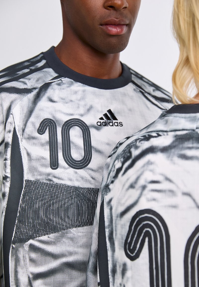 Vista parcial de dos personas vestidas con camisetas deportivas Adidas en blanco y negro con el número 10, enfrentándose entre sí contra un fondo liso.