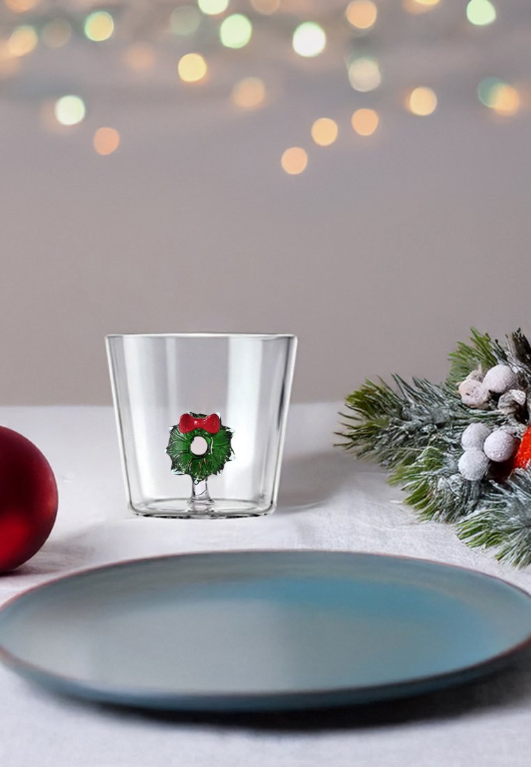 Przezroczysta szklanka z zielonym wzorem wianuszka z czerwoną kokardą. W pobliżu znajduje się niebieski talerz i czerwona ozdoba, a w tle widać zieleń.
