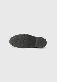 Semelle de chaussure en caoutchouc noir avec surface texturée, présentant des motifs ondulés et des grips circulaires pour l'adhérence. Pas de marque ni d'accents visibles.