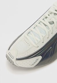 Witte en marineblauwe sneaker met een gestructureerde bovenkant, mesh panelen en vetersluiting; glanzende accenten aan de zijkant en geperforeerde details.