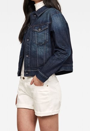 Kvinde iført en mørkeblå cowboyjakke og hvide shorts med venstre hånd i lommen, vist fra skuldre til midt på låret.