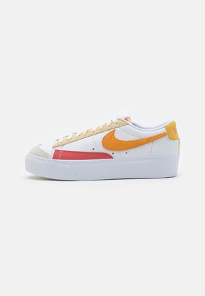 Baskets basses blanches avec une semelle épaisse, un swoosh Nike orange, des accents rouges et beige, et une languette de talon jaune sur un fond uni.