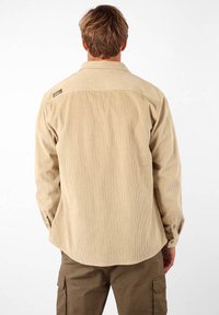 Chemise en velours côtelé beige clair avec un empiècement au dos, texture en côtes et poignets à boutons. Présente une petite étiquette de la marque sur l'épaule.