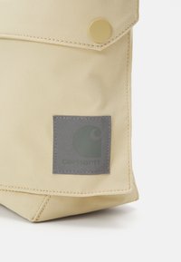 Beige szövet táska, fedelezett záródással, sárga gombbal és szürke, kerek mintázatú logóval. Látható varrások.