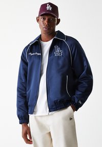 Navy athletic jacket with white piping and a logo, zippered front, elastic cuffs, paired with a maroon cap featuring a logo. White T-shirt underneath.