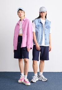 Deux personnes se tiennent côte à côte. L'une porte une chemise en velours côtelé rose, un crop top blanc, un short bleu marine et des baskets roses. L'autre a un gilet en denim, un t-shirt blanc, un short bleu marine et des baskets grises.