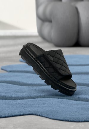 Juoda siūta šlepetė su storu gofruotu padu mėlynoje tekstūruotoje paviršiuje, fone neryški didelė pilka skulptūra.