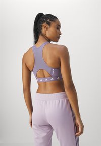 Lilla atletisk sportsbra med racerback-design, utskjæringsdetalj og logobånd. Kombinert med matchende lyse lilla bukser.