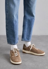 Sneaker in suede marrone con suola e lacci bianchi, caratterizzati da una texture trapuntata e dettagli dorati, indossati con jeans blu arrotolati e calzini bianchi.