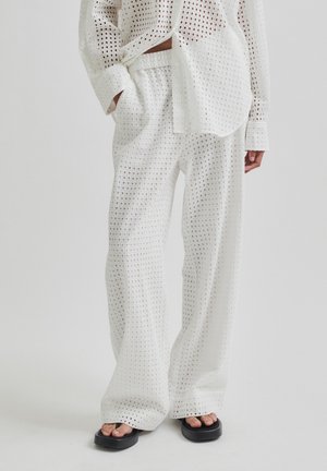 Chemise blanche à manches longues perforée et pantalon à jambes larges portés avec des sandales à plateforme noires, debout devant un fond uni.