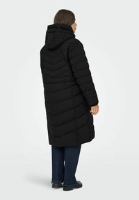 Abrigo acolchado negro con un diseño en forma de chevron, que incluye capucha, mangas largas y longitud por debajo de la rodilla. Lleva puesto sobre unos vaqueros azul oscuro.