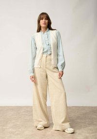 Biała kamizelka nałożona na jasnoniebieską bluzkę z falbanami, w zestawie z szerokimi beżowymi spodniami w paski i jasnymi trampkami. Proste, teksturowane materiały.