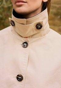 Beige trench coat magas gallérral, két nagy fekete gombbal és sima, matt felülettel. Varrott részletek az illesztések mentén.