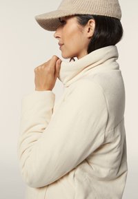 Chaqueta de pana beige con un cuello alto, que presenta una textura acanalada y un cierre de cremallera. Un gorro de punto beige complementa el look.