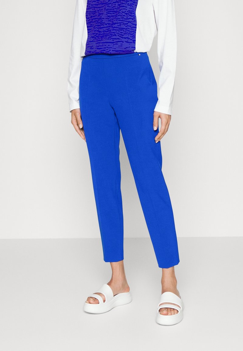 Femme portant un pantalon bleu vif ajusté, une chemise blanche à manches longues, un haut bleu à motifs et des sandales compensées blanches, debout sur un sol gris.
