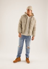 Veste matelassée en velours côtelé beige avec une capuche, portée sur une chemise claire, associée à un jean bleu déchiré et des bottes marron avec des accents jaunes.