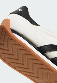 Basket blanche avec des bandes noires et languette au talon, semelle en caoutchouc marron en zigzag et logo Adidas partiel sur la languette du talon.