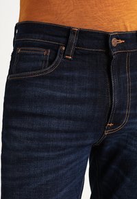 Mörkblå denimjeans med orange sömmar, en klassisk femfickdesign och en knappknäppning i midjan. Slät textur överlag.