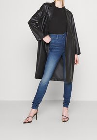 Cappotto lungo in ecopelle nera sopra un top nero aderente, abbinato a jeans skinny blu e tacchi alti neri con fasce. Texture liscia e vestibilità moderna.