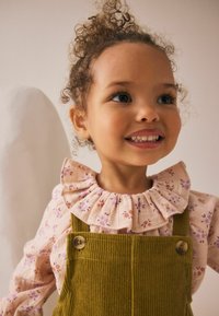 Das Kind trägt ein hellrosa, blumengemustertes Hemd mit einem gerafften Ausschnitt, kombiniert mit olivgrünen Corduroy-Overalls, die mit Knopfakzenten versehen sind.