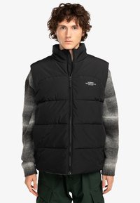 Svart vadderad väst med hög krage, framzipper och logodetalj. Bärs över en grå mönstrad tröja och kombineras med gröna cargobyxor.