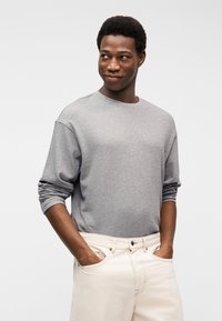 Νεαρός με κοντά σγουρά μαλλιά, που φορά γκρι πουκάμισο μακριάς μανίκας και κρεμ παντελόνι, με τα χέρια στις τσέπες, κοιτάζει πλευρικά με ένα ελαφρύ χαμόγελο.