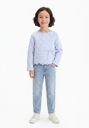 Niño con cabello hasta los hombros que lleva una camiseta acolchada de manga larga azul claro atada al costado, vaqueros azul claro y zapatillas blancas, sonriendo.