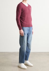 Pull bordeaux à col en V superposé sur une chemise blanche, associé à un jean denim bleu clair et des baskets blanches avec des détails. Texture douce et tricotée.