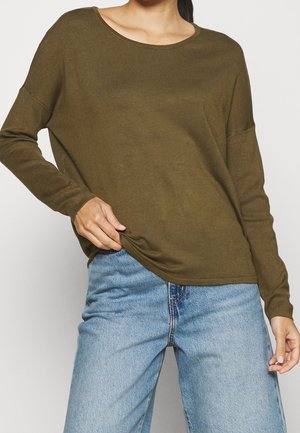 Kvinde iført olivengrøn langærmet top og lyseblå jeans, med den ene hånd trækker i toppen nederst.