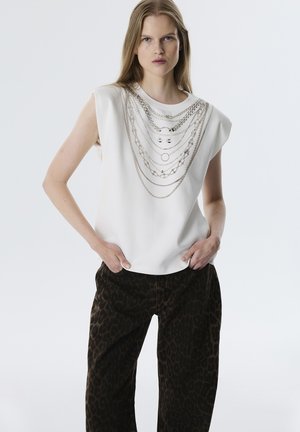 Witte mouwloze top met gelaagde metalen kettingontwerp bij de halslijn, gecombineerd met donkerbruine broek met luipaardprint en wijde pijpen.