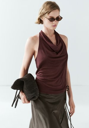 Frau trägt dunkle Sonnenbrille, ärmelloses bordeauxfarbenes Top, olivfarbigen Rock und hält eine gefaltete dunkle gewebte Tasche in ihrer linken Hand.