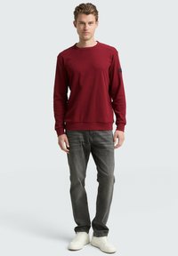 Felpa bordeaux con finitura testurizzata, scollo rotondo e polsini a costine, abbinata a jeans grigio scuro e sneakers bianche.