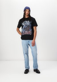 Černé bavlněné tričko s grafikou Star Wars, kombinované se světle modrými džíny a černými botami. Model má na sobě modrou čepici.