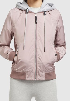 Personne portant une veste bomber rose clair zippée avec un sweat à capuche gris en dessous, mains dans les poches de la veste, pantalon gris.