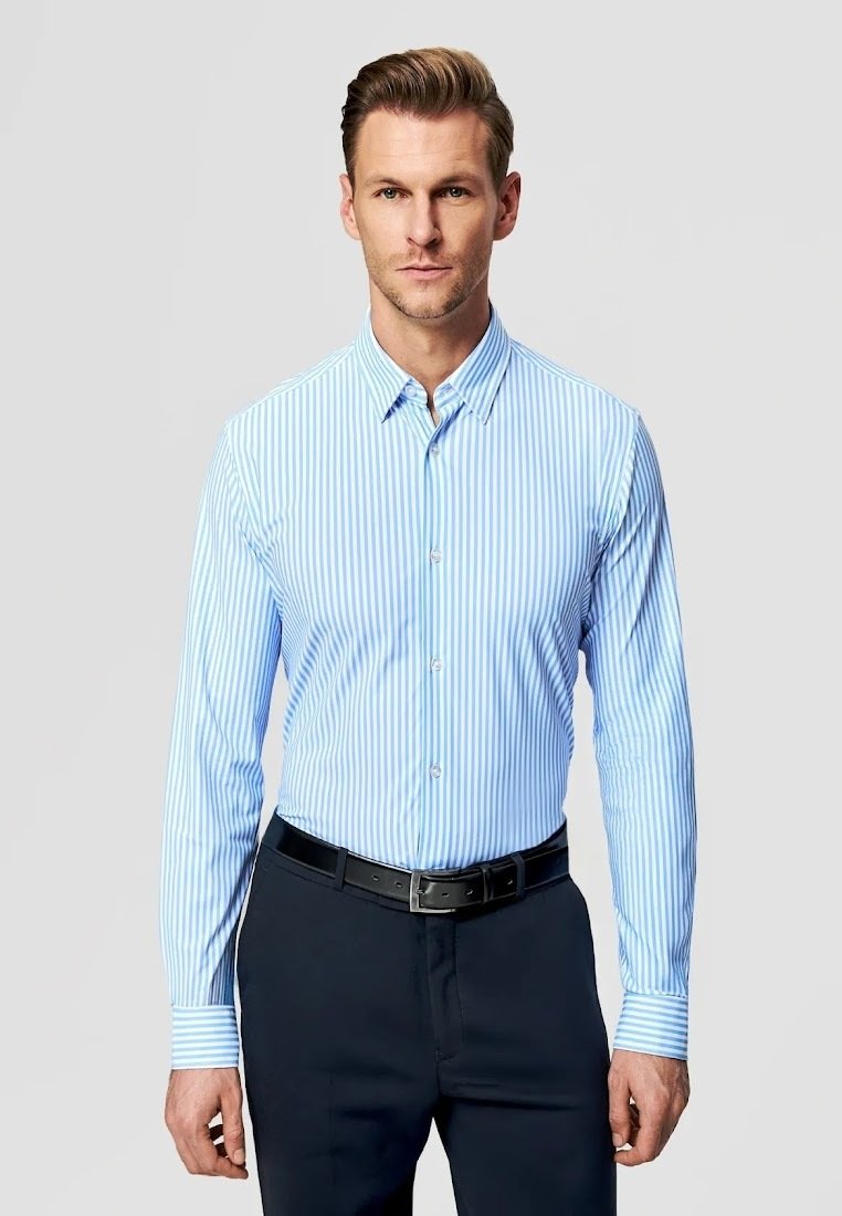 Mężczyzna w jasnoniebieskiej i białej koszuli w paski z długim rękawem, włożonej do ciemnych spodni z czarnym paskiem, stojący na jednolitym tle.