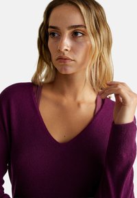 Sweter w serek w głębokim purpurze, wykonany z miękkiego materiału. Długie rękawy z prążkowanymi mankietami. Nałożony na brązowy top na ramiączkach.