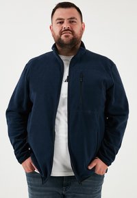 Tamsiai mėlynas fliso džemperis su aukštu apykakle, pilno ilgio užtrauktuku ir dviem šoninėmis kišenėmis. Yra krūtinės kišenė su užtrauktuku.