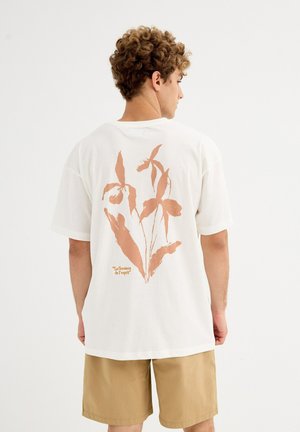 Joven con cabello rizado que lleva una camiseta blanca con un gran estampado botánico marrón y texto, combinada con pantalones cortos color beige, de espaldas.