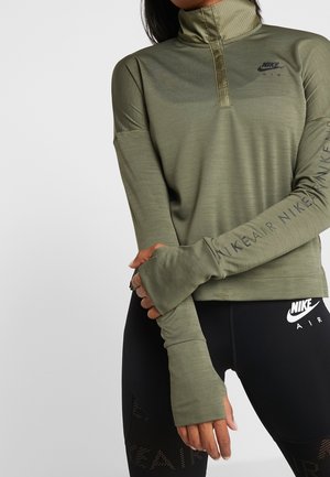 Femme portant un haut Nike Air à manches longues vert olive avec des trous pour les pouces et un legging noir Nike Air, bras croisés à la taille.