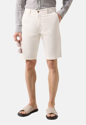 Man draagt witte op maat gemaakte shorts, grijs overhemd met lange mouwen, beige slippers, houdt zonnebril in linkerhand, rechterhand in zak.