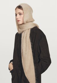 Beige, flauschiger Wollschal, der als Kapuze und Wickel getragen wird. Weiche Textur, lang, legt sich um den Hals, kombiniert mit einem dunklen Wildledermantel.