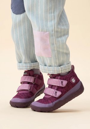 Chaussures d'enfants violettes et roses avec des straps en velcro, en tissu texturé et une semelle en caoutchouc solide. Portées avec un pantalon rayé bleu clair avec des patchs.