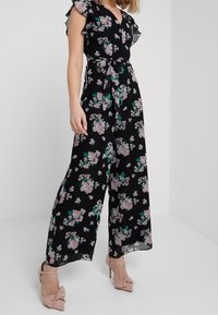 Černý květinový overal s krátkými volány, vázaným pasem a širokými nohavicemi s růžovými a zelenými květinovými vzory. Spárováno s světle růžovými podpatky.