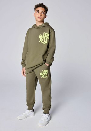 Teenager trägt olivgrünen Hoodie und passende Jogginghose mit neon-gelbem "AllSaints London" Logo, steht mit einer Hand in der Tasche.