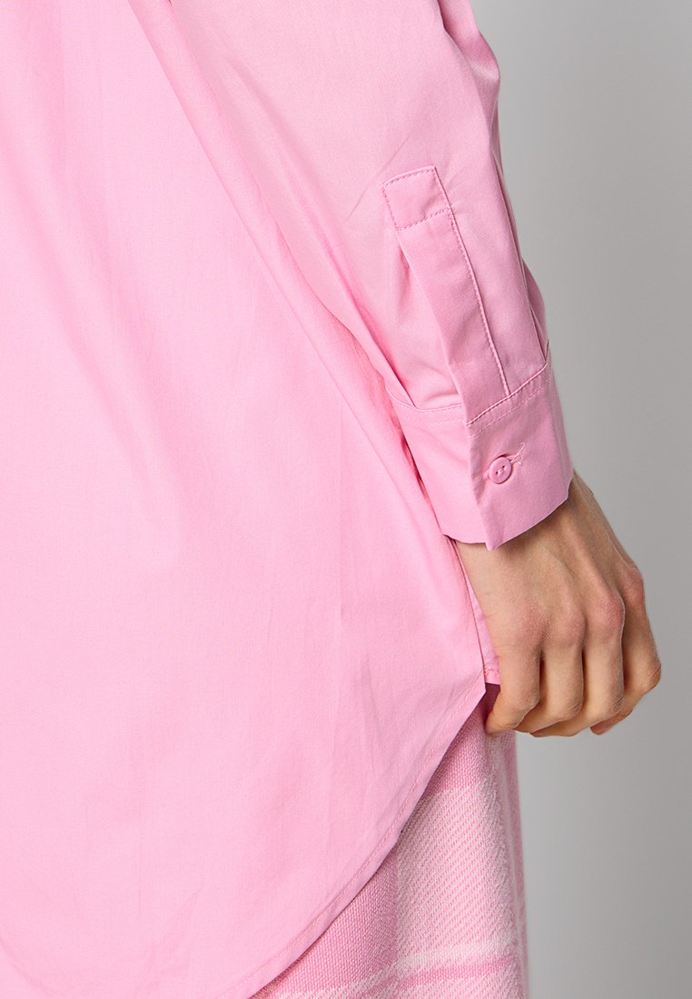 Chemise rose à manches longues en coton. Présente un ourlet arrondi, des poignets boutonnés et une texture lisse. Une main tenant l'ourlet est visible.